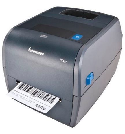 A gray Honeywell PC43T barcode label printer is printing a label. The printer is positioned on a white background, and it has a label feeder on the left side from which labels are dispensed.