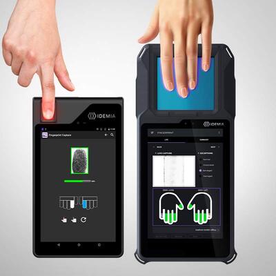 An IDEMIA biometric tablet being used for fingerprint enrollment, with a hand placing their finger on the device's screen. The tablet has a black casing and a display showing the biometric interface.