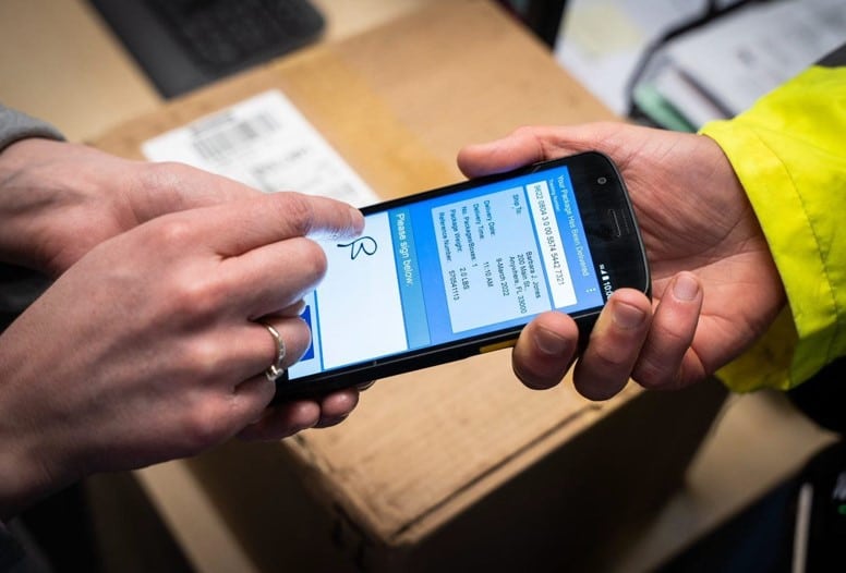 A person holding a Zebra TC22 mobile computer, displaying a screen with inventory management software.